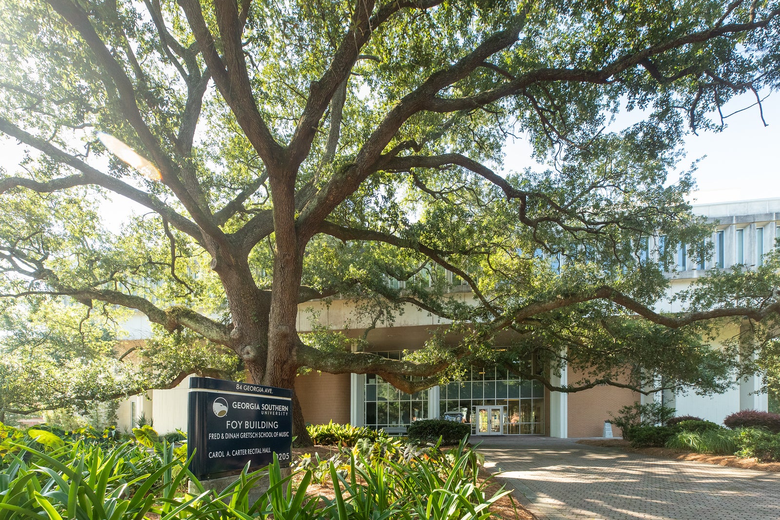 A brick building slightly hidden amongst sweeping green trees framed by a blue sky. There is a blue sign in front of the building that says "Foy Building."