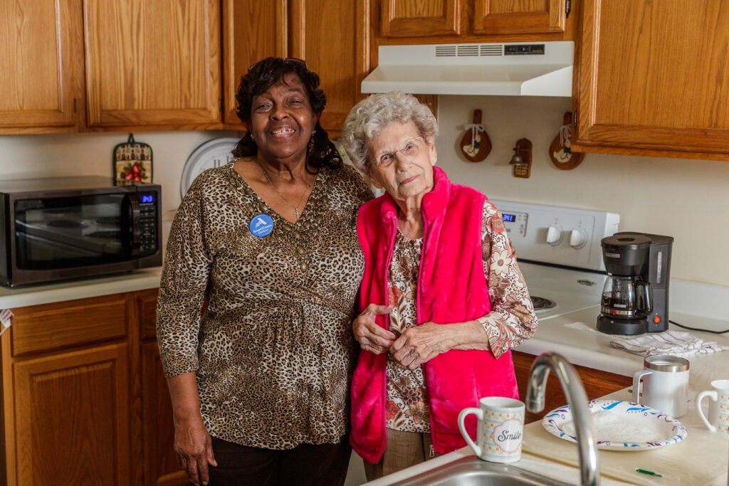 Two women stand together in a warm kitchen, smiling at the camera; one wears leopard print, the other a bright pink vest.