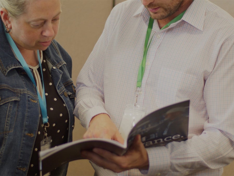 Two adults review a brochure together at a conference; man in a white checking shirt with a green lanyard, woman in a denim jacket with a blue lanyard.