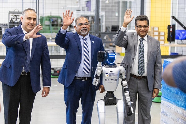Three people are standing in a lab with a humanoid robot. Everyone is waving.