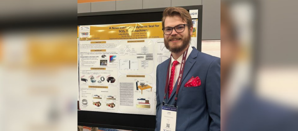 Man with glasses and a beard in a blue suit and red tie stands beside an academic poster at a conference. This is an informative image of a presenter with a poster.
