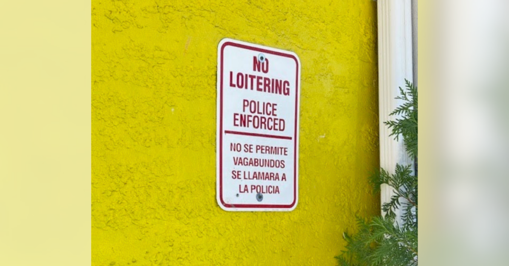 No loitering sign—Police Enforced, bilingual English and Spanish, mounted on a bright yellow textured wall.