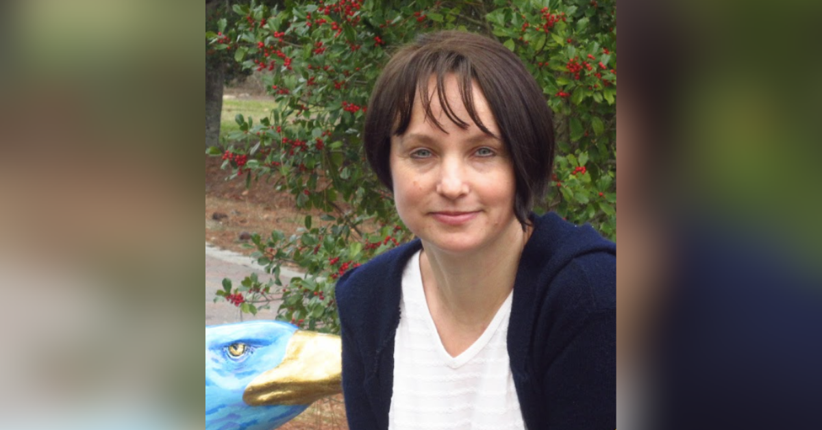 Woman with short dark hair and a white top, smiling outdoors with red-berry bushes behind her.