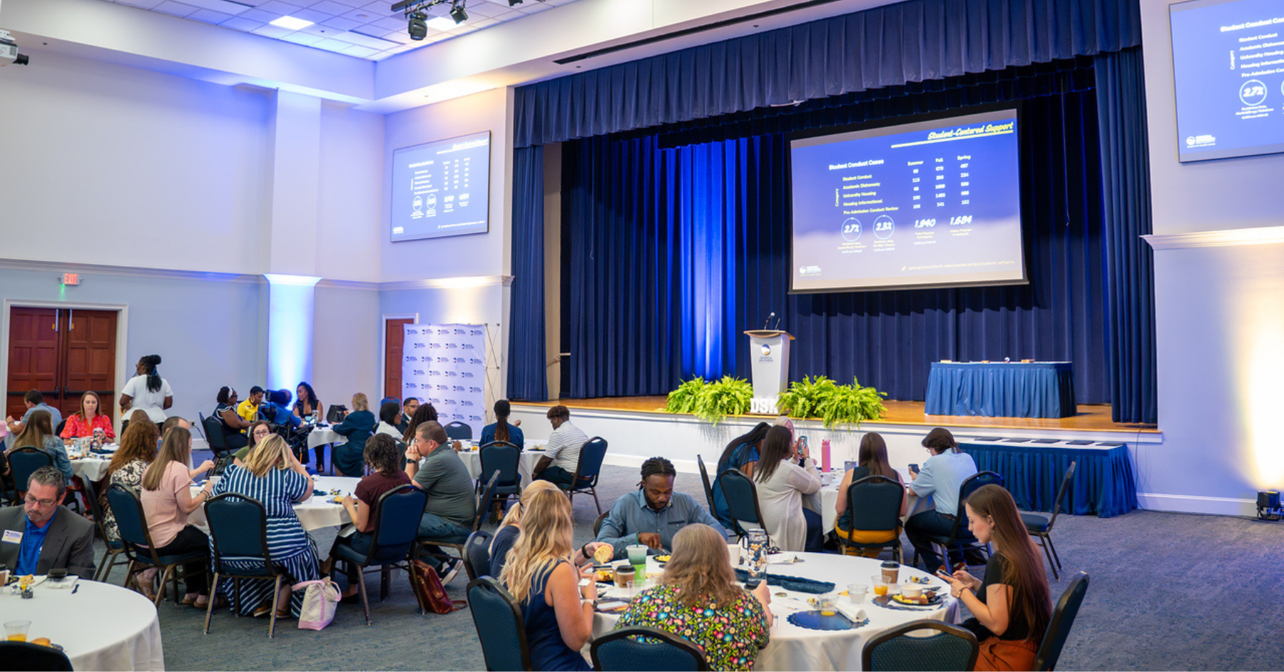 Conference hall with round tables and guests dining, stage with podium and large screens displaying slides on a blue-lit backdrop.