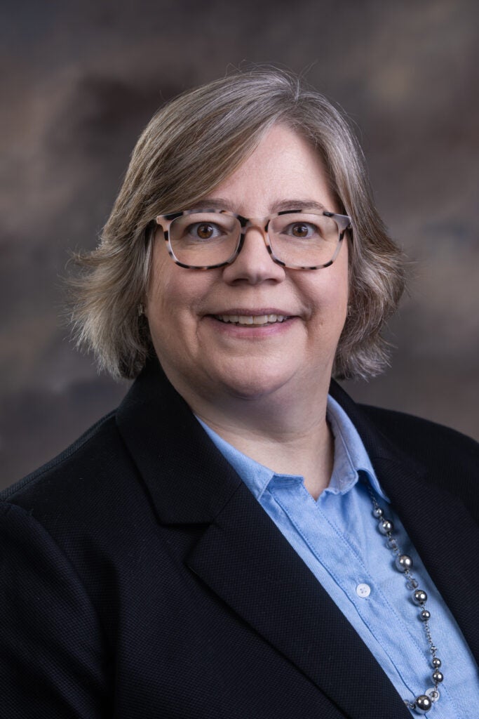 Image is a professional headshot of a smiling person with glasses, a blue shirt, and black jacket. 