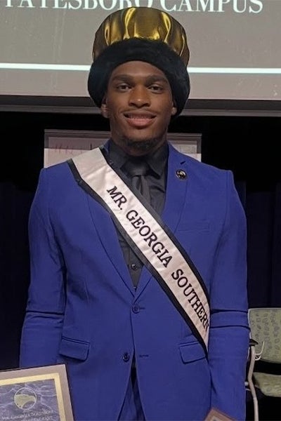Man on stage wearing a royal blue suit, black shirt and tie, a black-and-gold hat, and a sash reading 'MR. GEORGIA SOUTHERN'.