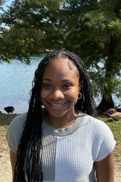 Young woman with braided hair wearing a light sweater, smiling at the camera with a lake and trees in the background.