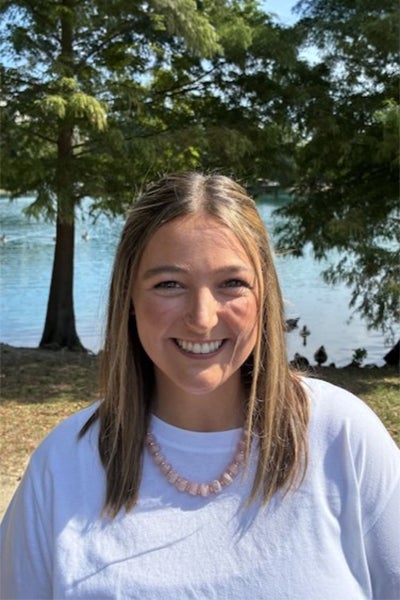Smiling woman outdoors by a lake with trees in the background, wearing a white shirt and pink bead necklace.