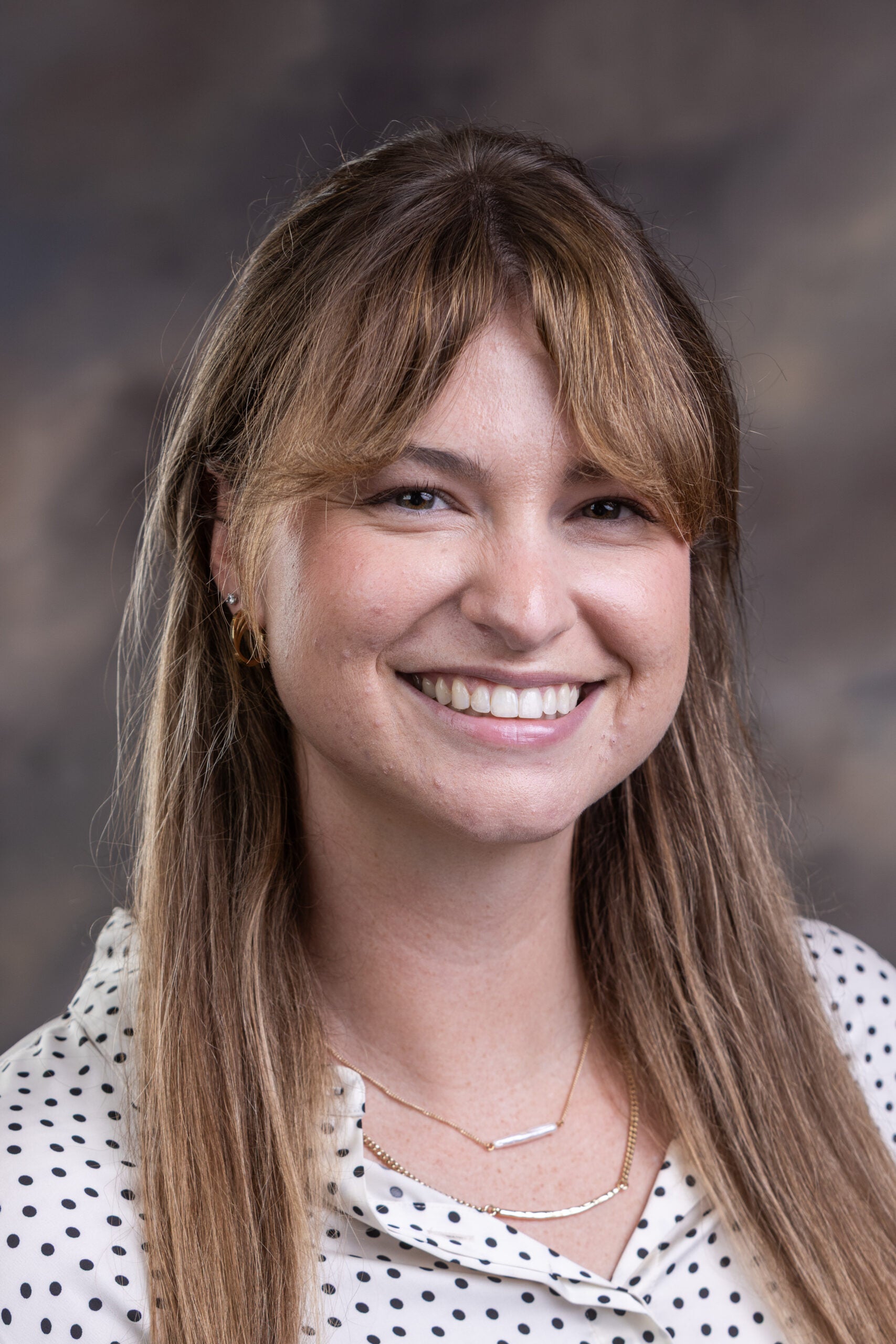 Portrait of a smiling woman with long light brown hair, wearing a white polka-dot blouse and layered gold necklaces, neutral background.