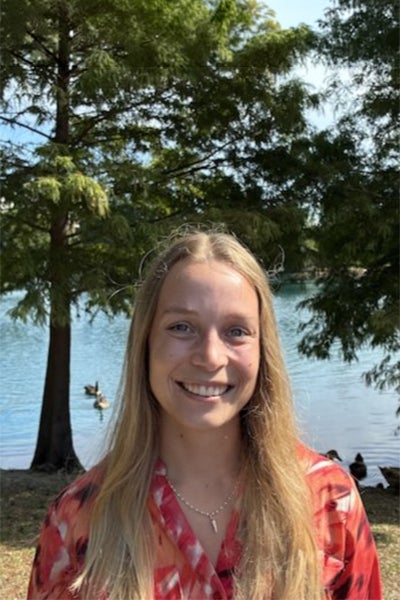 Smiling woman with long blonde hair wearing a red shirt, standing by a lake with trees in the background.