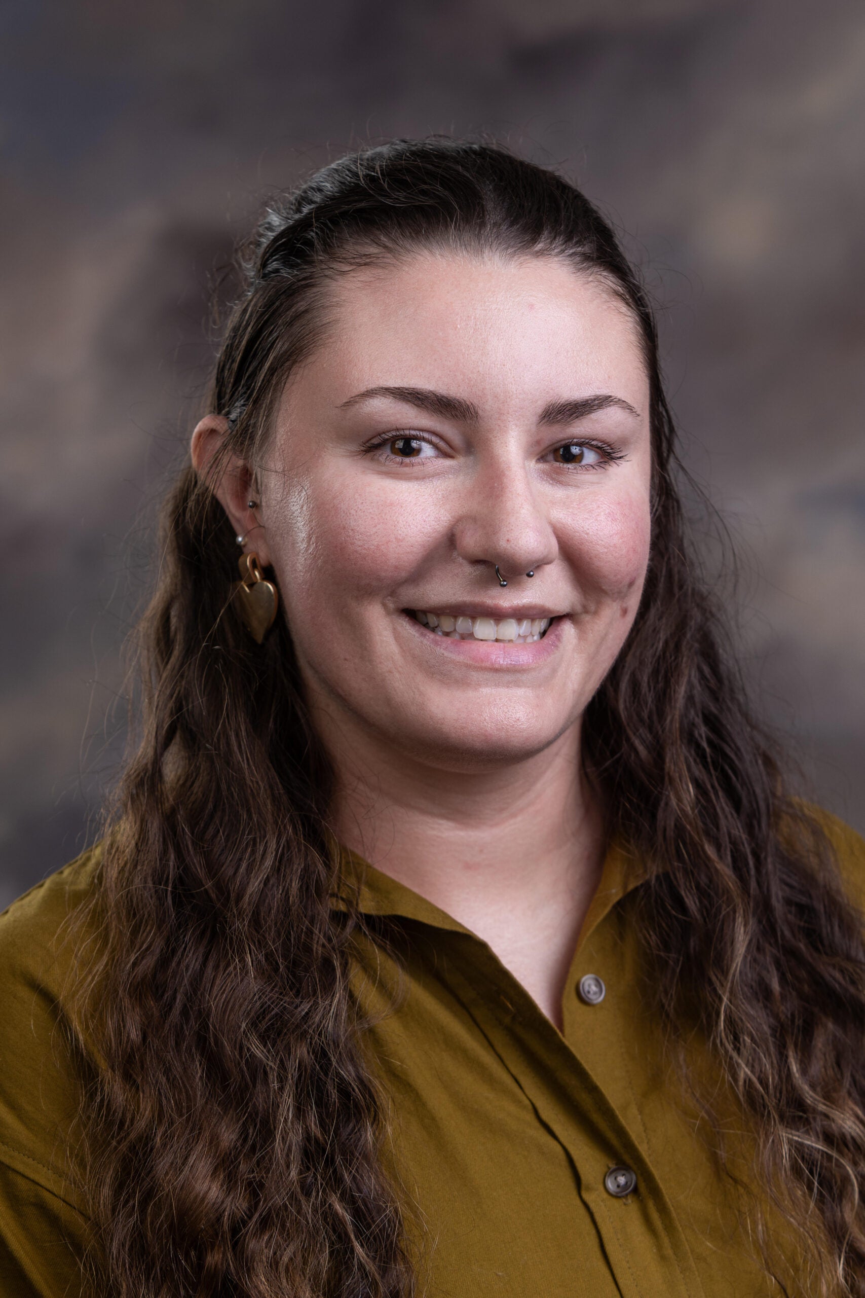 Smiling young woman with long wavy brown hair, wearing an olive green button-up shirt, head-and-shoulders portrait.