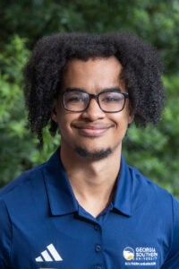 Portrait of a smiling man with curly hair and glasses wearing a blue Georgia Southern University polo outdoors.