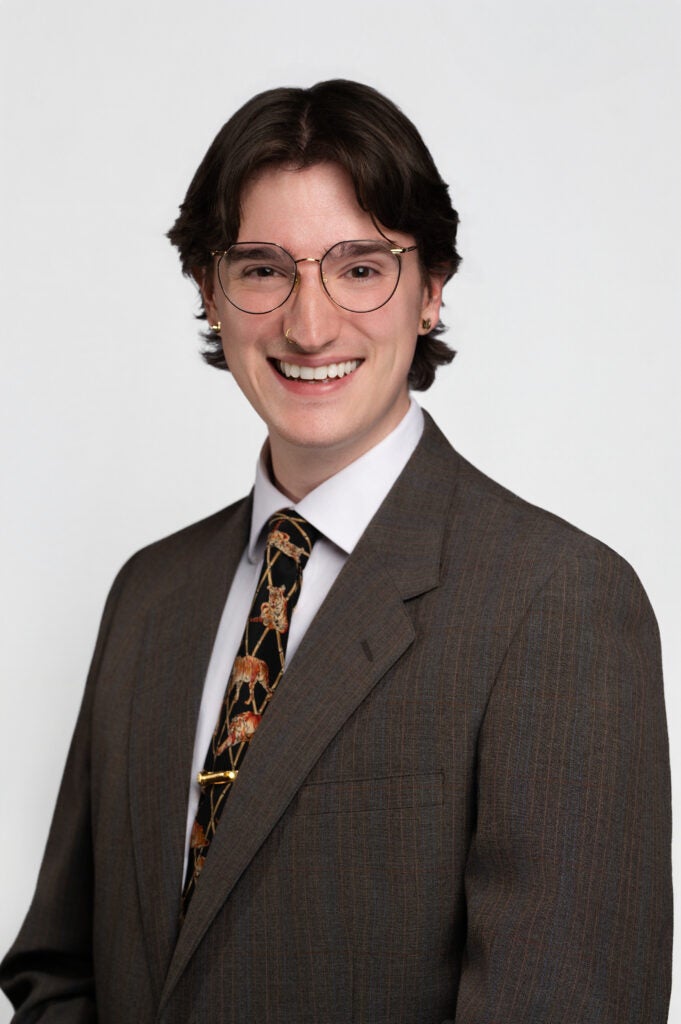 Smiling person in a brown pinstripe suit, white shirt, patterned tie, round glasses, against a light background.