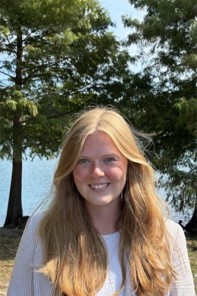 Young woman with long blonde hair and a light top smiling outdoors by a lake with trees in the background.
