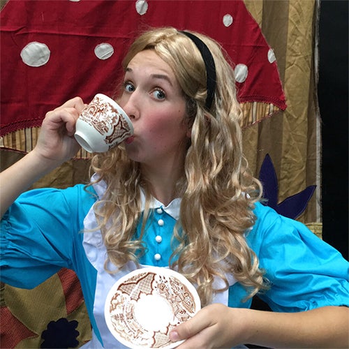 Young woman in a bright blue period costume drinks from a decorative teacup on a theater stage, holding a matching plate behind her.