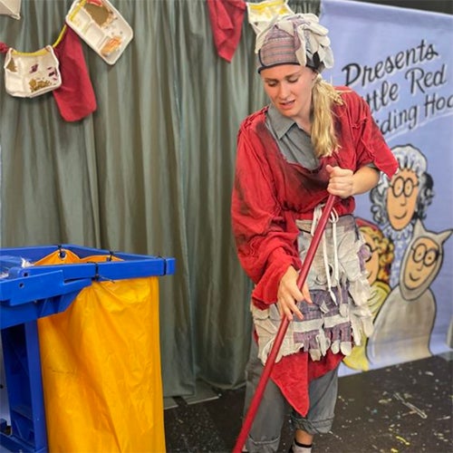Person dressed as Little Red Riding Hood sweeping the floor with a broom on a stage set, with a blue recycling bin and a cartoon backdrop.