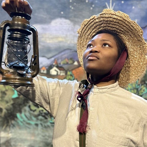 Person in a wide straw hat holds up an old lantern, looking upward, with a painted mural backdrop suggesting a historical or storytelling exhibit.