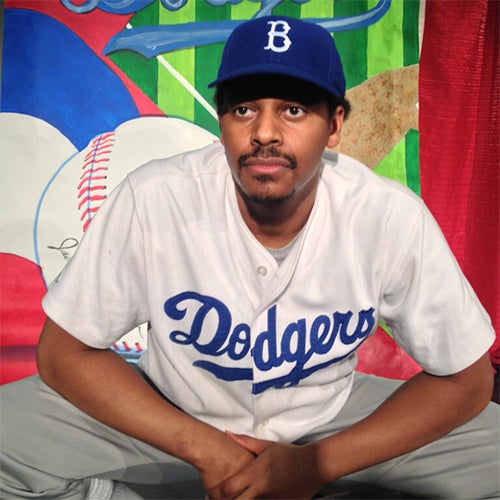Portrait of a man wearing a Dodgers jersey and blue cap, seated with hands folded, against a bright mural background.