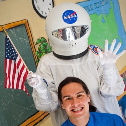 Person in a white astronaut suit and NASA helmet posing with an American flag, smiling with a student in front in a classroom set.