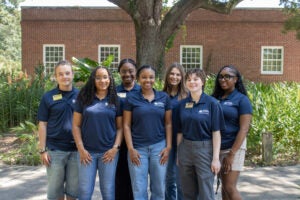 Flight Leaders on the Armstrong Campus stand for a group photo outdoors. Trees and shrubbery are behind them and a brick building is in the background. The Flight Leaders are all wearing navy blue polo shirts.