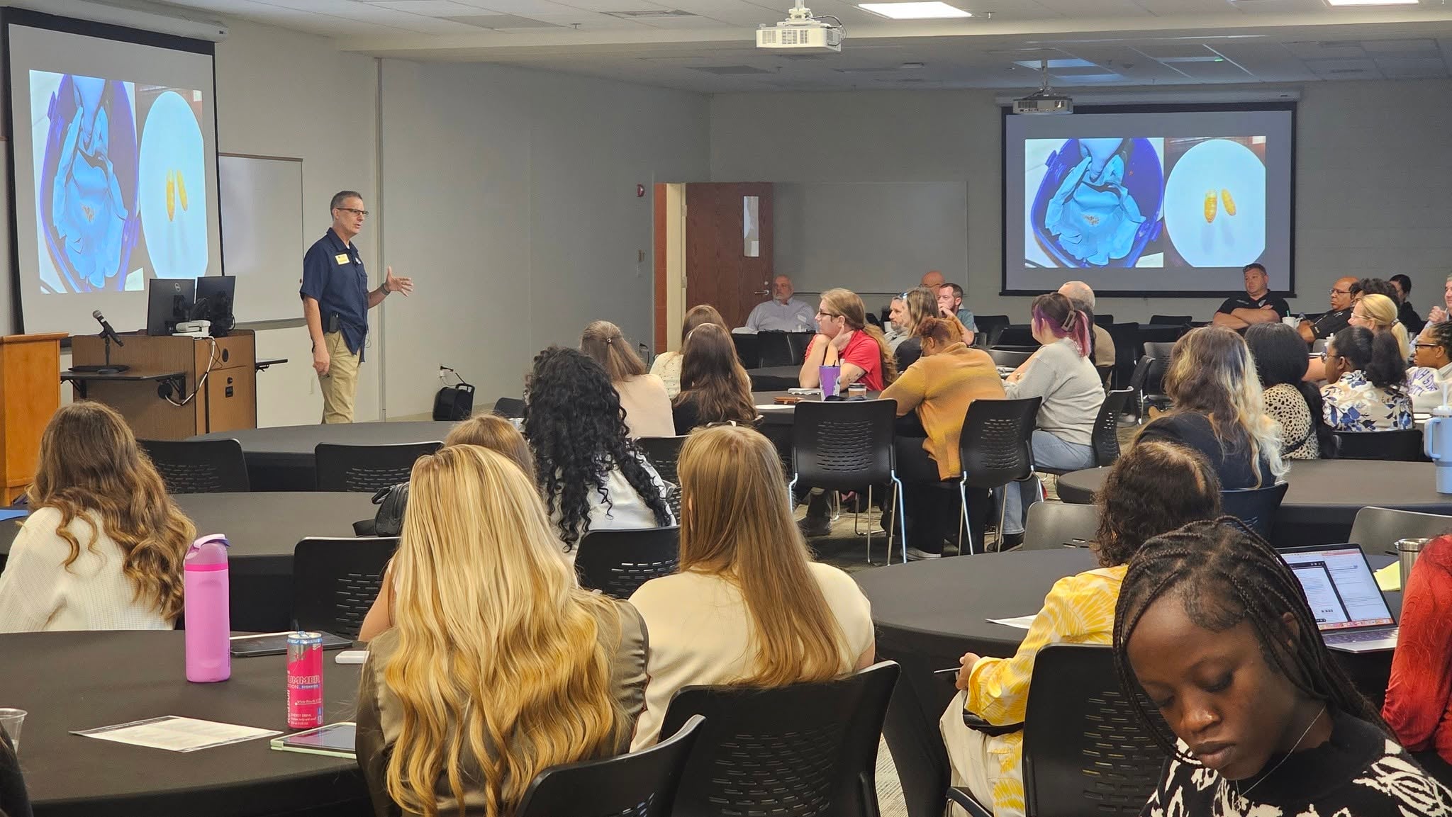 Professor Ed Mondor stands in front of a large room. An audience of a few dozen people are seated at tables around the room. A projector screen shows an image of a medical scan.