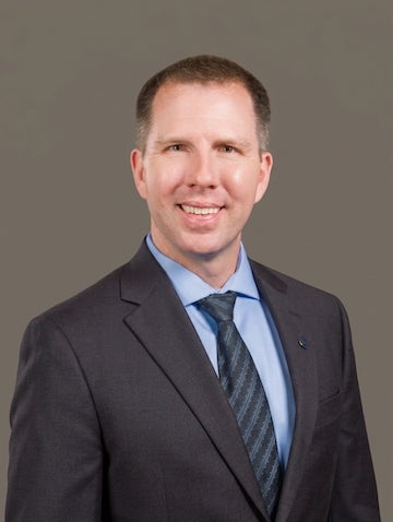 Someone poses in front of a gray backdrop. They are wearing a dark gray suit jacket, blue shirt, and gray striped tie. 