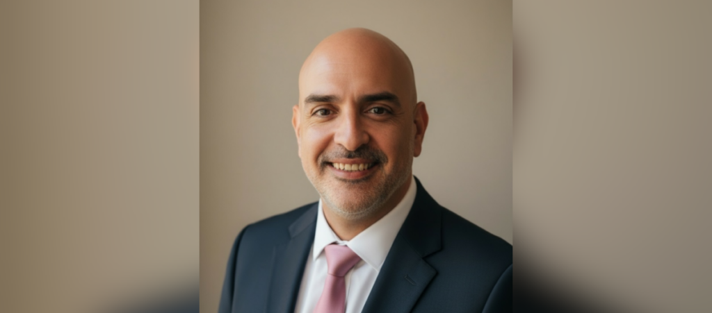 Dr. Francisco Lugo wears a blue-ish black suit with a white collared shirt and pink tie in a portrait photo against a beige background.