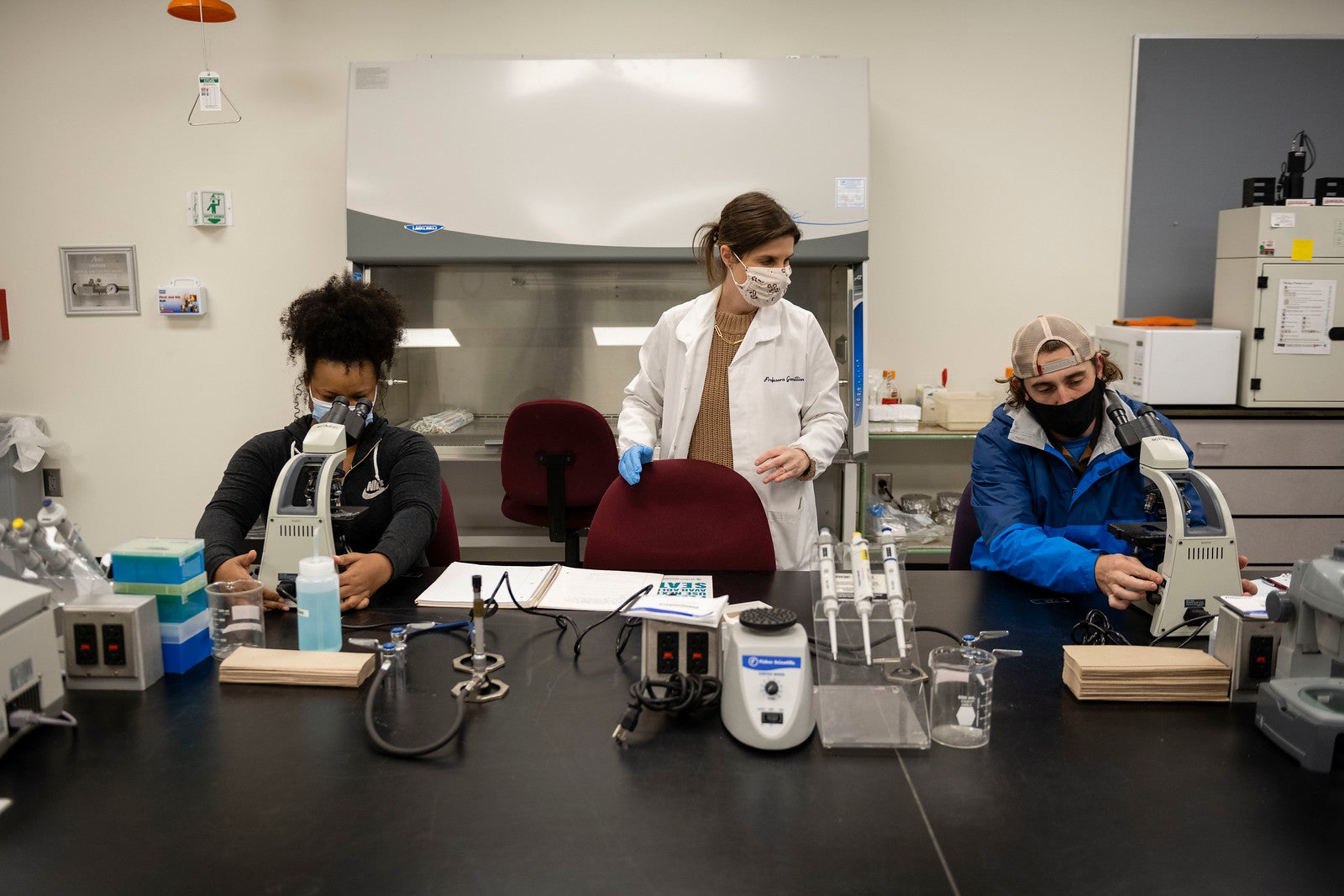 Dr. Sara Gremillion wears a white lab coat and a face mask while working with students who are using microscopes in a lab. She is the center of the photo with one student to her left and one to her right.