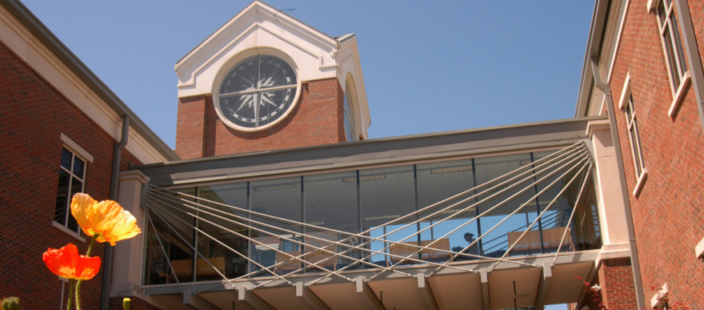 The Science Center of the Armstrong Campus of Georgia Southern is behind some orange flowers and in front of a blue sky. There is a glass walkway connecting two brick buildings.