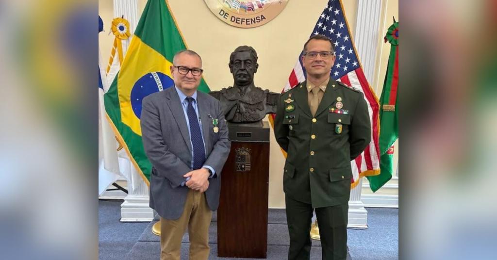 Two men stand in front of a bronze bust with the Brazilian Flag and American Flag in the background. The man on the left wears a grey suit and dons the Brazilian Army Medal, and the man on the right wears a green, formal military uniform with several medals and ribbons attached around the chest. 